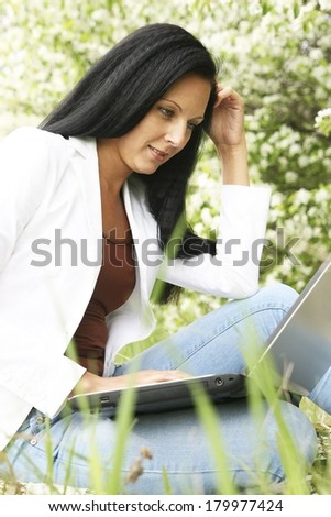 Portrait of Young adult beautiful brunette girls working on laptop at summer or spring  park on green fresh grass Woman looking at computer screen typing on the keyboard and thinks about something