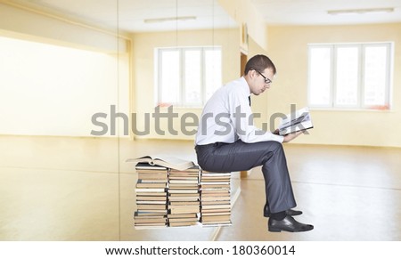 Businessman sitting on a stack of books and reading opened big book inside in empty room with window
