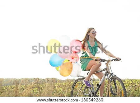 Copy space for inscription SYMBOL OF EASE Young Girl in sexy dress with sunglasses riding bicycle flying air balloons on leash yellow sun set sky background