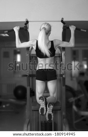 Back view Fitness Woman with smiling face. Close-up young adult blond girl with long hair holding simulator for swing press on dark green wall background