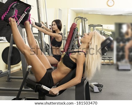 Portrait of two young adult Girls do exercise for legs and hands. in fitness gym on mirror with reflection and window background 2 woman with long blond and brunette hair sitting smiling face