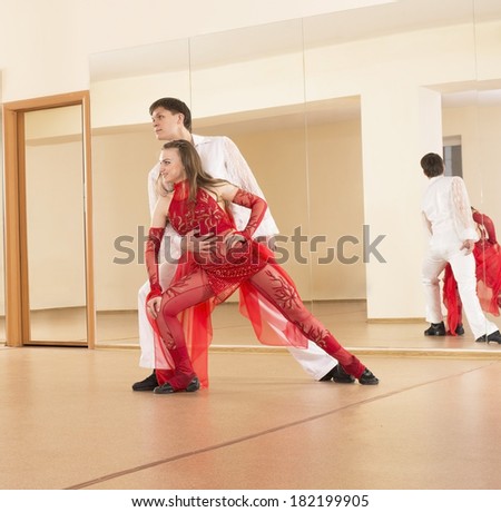 Portrait of couple young adult Latino dancers in action Woman in red transparent dress and caucasian man in white shirt and trousers embracing inside or indoor on mirror yellow wall background