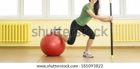 Smiling sporty slim sexy woman in sporty uniform crouches with fit bar and based leg on red rubbish fitball on yellow wall and windows background