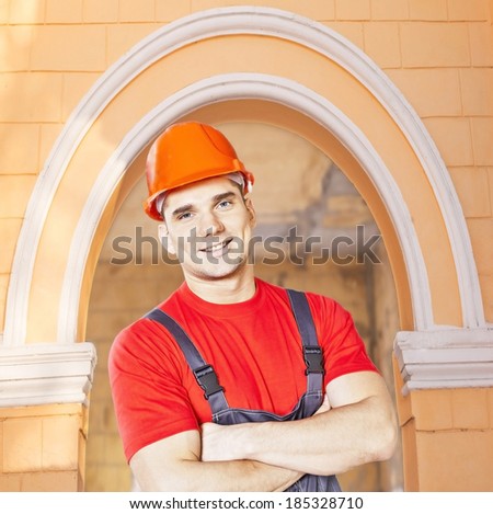 Portrait of young adult worker in blue and red uniform and security orange helmet standing above green arch building in perspective background