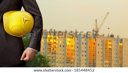 torso engineer hand  holding yellow helmet for workers security on background of new highrise apartment buildings window construction cranes on background of evening sunset cloudy sky Crane lifts load