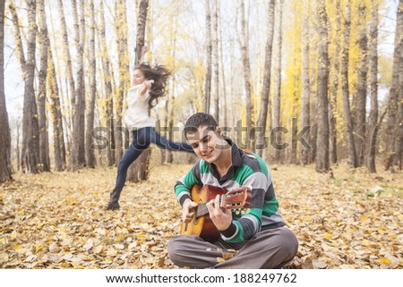 Portrait of cute brunette man sit at fall  leaves playing on acoustic guitar and latin hispanic girl is running and dancing around on yellow autumn forest background  in park