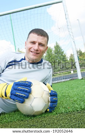Portrait of young adult Soccer football goalkeeper lie on fresh green grass on gate net and blue cloudy sky background Goalkeeper looking at camera