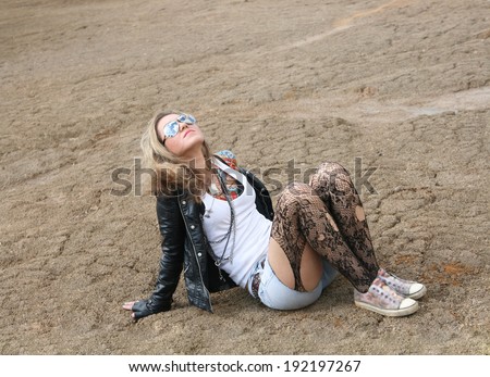 sexy Freak woman painted body art tattoo wearing leather jacket, sunglasses and white shirt, jeans shorts and torn pantyhose on the background of empty desert land Freaky young adult girl looking up