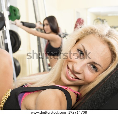 Portrait of two young adult Girls do exercise for legs and hands. in fitness gym on mirror with reflection and window background 2 woman with long blond and brunette hair sitting smiling face