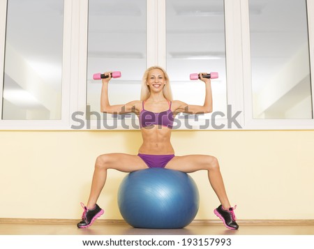 Portrait of Sweet blond very hot sexy slim young adult girl doing fitness exercises sitting on blue rubber ball  with dumbbells agains yellow wall and windows background