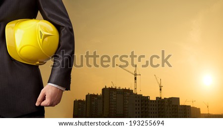 engineer holding yellow helmet for workers security on background of new highrise apartment buildings and construction cranes on background of evening sunset cloudy sky Silhouette Crane lifts load