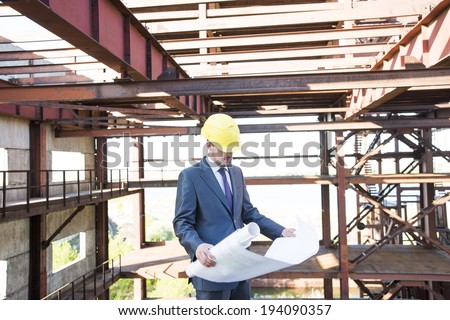 picture of young adult man engineer holding some blueprints and his hand in his pocket and looking at paper plan. on brown metal beam construction building background in perspective
