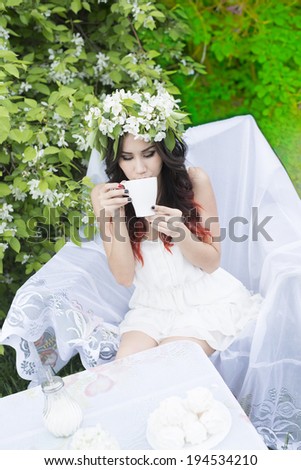 Portrait of beautiful latin hispanic girl sitting with bloom spring bouquet in white armchair Cute woman wearing sexy dress has a rest in summer park cafe against blooming apple trees background