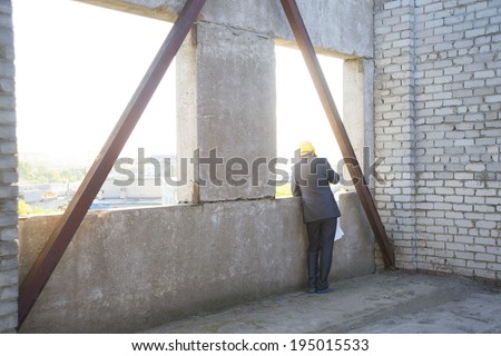 Back view engineer with helmet for workers security holding in hands blueprint paper plan, talking mobile phone and looking in window on background of  wall building  Unrecognizable person