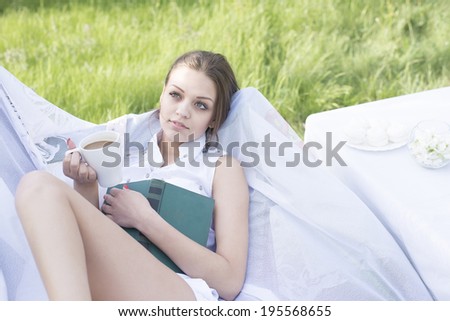 beautiful young adult caucasian girl laying with green book and has a rest in park cafe on fresh green grass and blooming tree background Slim female sit on white armchair and dreaming