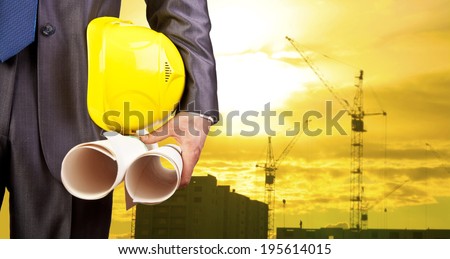 engineer\'s torso and hand holding yellow helmet for workers security on the background of a new highrise apartment buildings and construction cranes and evening sunset sky