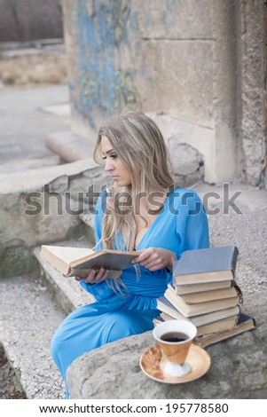 Portrait of sad dreaming  blond girl with long blond hair holding big old retro book near stack and cup of tea Slim sexy woman sitting on concrete ladder