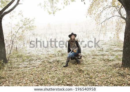 Young alone slim girl in a black leather jacket with fur collar sitting on the road in a forest in an old retro suitcase on snowy background of the autumn snowfall