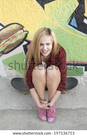 Portrait of beautiful teen girl sitting on skateboard over wall background with abstract graffiti art. Urban outdoors teenager\'s lifestyle Full length Cute young woman sit skate in denim jeans shorts