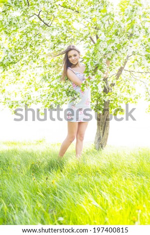 Portrait of young adult caucasian Girl holding branch with blooming spring flowers on apple tree background. Full length Cute female standing barefoot on fresh green grass