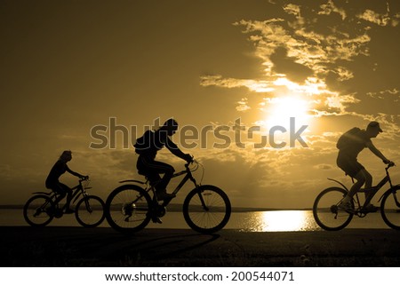 Image of sporty company three friends on bicycles outdoors against sunset. Silhouette motion of 3 cyclist along the shoreline coast and cloudy sunset sky background Space for inscription