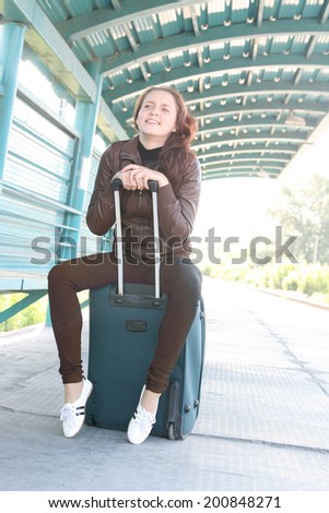 Full length Pretty young adult curly redhead woman with suitcase a metal wall in perspective at the station Beautiful girl wearing brown casual dress Copy space for inscription