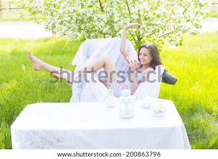 beautiful young girl sitting with cup of coffee and white sweat in park cafe near decor table on green grass and blooming tree background female sit on white armchair Copy space for inscription