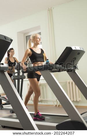 Vertical format Portrait of Two young adult cut sporty women run on machine in the gym centre Beautiful girls with white headphone inside on on wall and window background Copy space for inscription