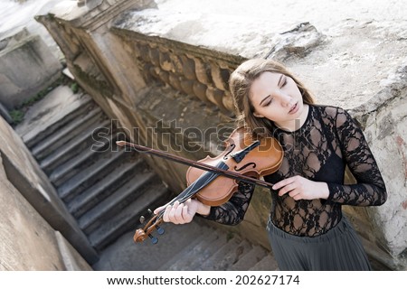 Portrait of Young adult sexy girl with long brown hair with a broken violin on old retro fence and concrete stairway in the Gothic style of classicism Cute slim woman wearint transparent black shirt