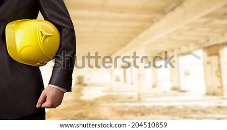 torso engineer hand holding yellow helmet for workers security against the support concrete beams of the unfinished industrial workshop or room inside Copy space for inscription