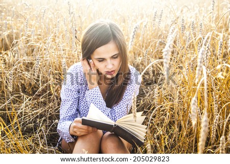 Portrait of young girl sitting on golden reap wheat in yellow sunset field Woman holding in hands black interesting book, reading text Empty Copy space for inscription Sun rays on brunette long hair