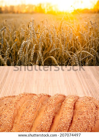 Fresh bread with seeds on wooden table over yellow wheat field backdrop of ripening ears of on sunset cloudy orange sky background of the setting sun on horizon