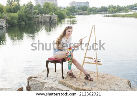 Young adult artist full length girl with body art on leg painting an summer landscape near clear water Woman sitting on stone mountain beach Empty copy space for inscription