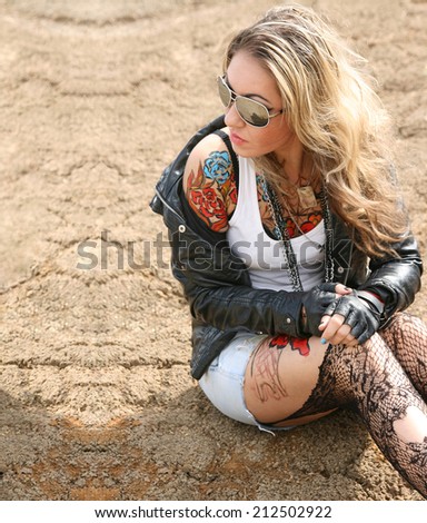 Portrait of stylish tattoo woman painted body art in the form of red blue flower in the white t-shirt, black leather jacket and denim shorts and torn pantyhose on background of gray desert