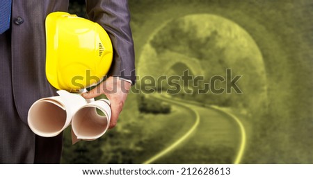 engineer or worker holding in hand helmet for workers security and blueprint paper plan on background of an underground mine with arc legs and rails for trolleys with coal in perspective Copy space