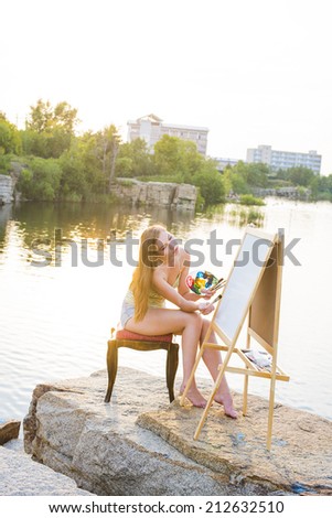 Portrait of Young adult artist full length girl with body art on leg painting an summer landscape near clear water Woman sitting on stone mountain beach Empty copy space for inscription