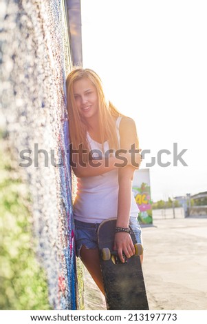 Portrait of beautiful smiling girl wearing white shirt stands with skateboard based on graffiti wall on sky background with sun set rays Empty copy space for inscription
