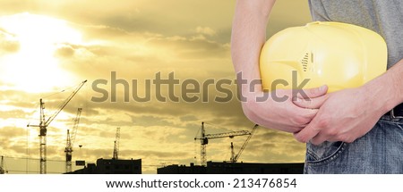 engineer or worker holding in hand yellow helmet for workers security on background of highrise apartment building, construction cranes on background of evening sunset sky Silhouette Crane lifts load