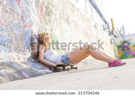 Portrait of beautiful teen girl sitting on skateboard over wall background with graffiti art. Urban outdoors, teenager's lifestyle. Full length Cute young adult woman sit skate in denim jeans shorts