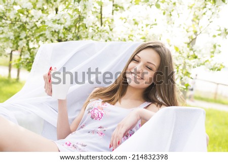 beautiful young adult caucasian girl laying has a rest in park with cafe on fresh green grass and blooming apple tree background Slim female sit on white armchair and laugh with closes eyes