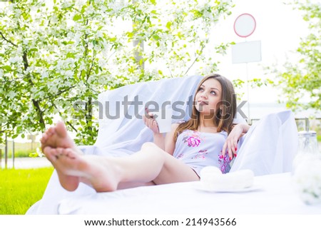 girl sitting with coffee cup and has a rest in spring park cafe on fresh grass, blooming apple tree background Slim woman sit on white armchair put your feet up on table and dreaming looking far away