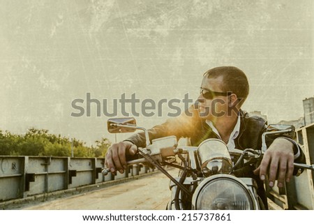 Romantic portrait handsome biker man in sunglasses sits on a bike on a sunset  on old rusty metal bridge in city against house and sunny sky background Empty copy space for inscription