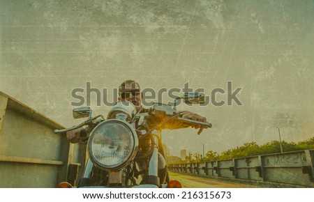 Romantic portrait handsome biker man in sunglasses sits on a bike on a sunset on old rusty metal bridge in city against house and sunny sky background Empty copy space for inscription