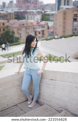 Young adult brunette girl in casual clothes, stand on edge of terrace, dressed in blue shirt and jeans , Cute woman with long hair looking at far on big city , flowers sky and bush background