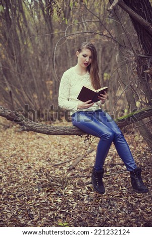 woman with long wet hair in the autumn forest reading big book  Girl sit on trunk in fall park and looking at page  Empty copy space for inscription Female wearing blue leather trousers under rain