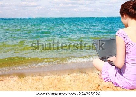 Back view of redhead girl using black laptop on yellow sand beach Young adult woman looking at far away on water in river texture and cloudy sky background Empty copy space for inscription