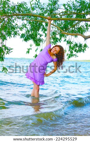 Beautiful young adult curly redhead woman standing in warm water in lake or river under tree and keep trunk Empty copy space for inscription Girl wearing very sexy hot  violet dress