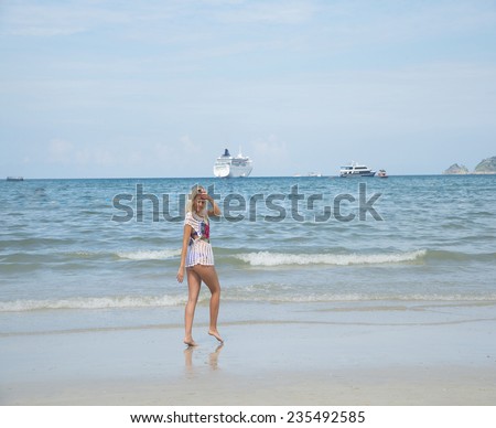 Bikini blond model standing dancing at shallow water with cruise liner,yacht, boat on background blue sky with clouds Cute woman or blondy girl with sexy legs wearing white transparent knitted tunic