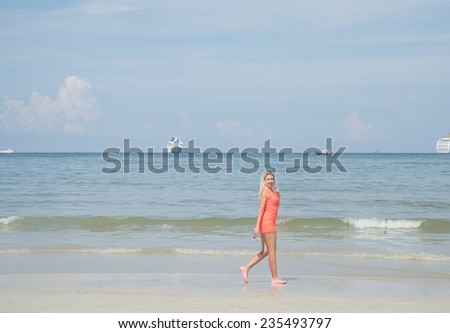 Bikini blond model standing dancing at shallow water with cruise liner,yacht, boat on background blue sky with clouds Cute woman or blondy girl with sexy legs wearing short red dress and sunglasses