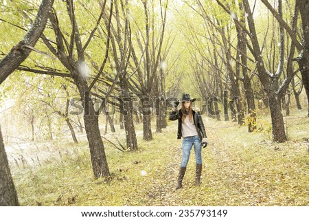 Outdoors portrait of casual clothed gorgeous fall fashion girl on green yellow autumn snowy park background Cute slim woman stand between cold tree and looking at camera Female wearing cowgirl hat